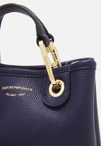 Borsa a mano in pelle blu navy con finitura testurizzata, hardware dorato e un logo prominente. Dotata di un accento metallico rotondo e di un dettaglio di chiusura sicuro.