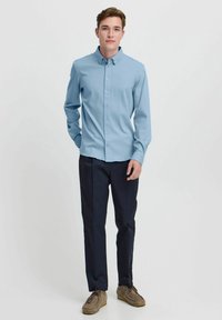 Camisa azul clara de botones y mangas largas, combinada con pantalones azul marino oscuros y zapatos marrones, frente a un fondo blanco liso.