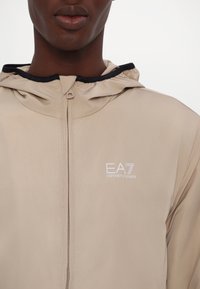 Beige Zip-Up-Hoodiejacke aus einem glatten, leichten Stoff. Mit schwarzem Besatz entlang der Kapuze und einem weißen gestickten Logo auf der Brust.