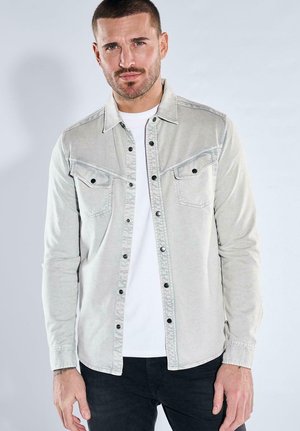 Mann mit kurzen Haaren und Bart, der ein hellgraues, geknöpftes Hemd über einem weißen T-Shirt und schwarze Hose trägt, vor einem schlichten Hintergrund stehend.