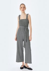 Svart och vit randig jumpsuit med vida ben, rynkad kroppdel och tjocka axelremmar. Inkluderar ett tygbälte för justerbar passform.