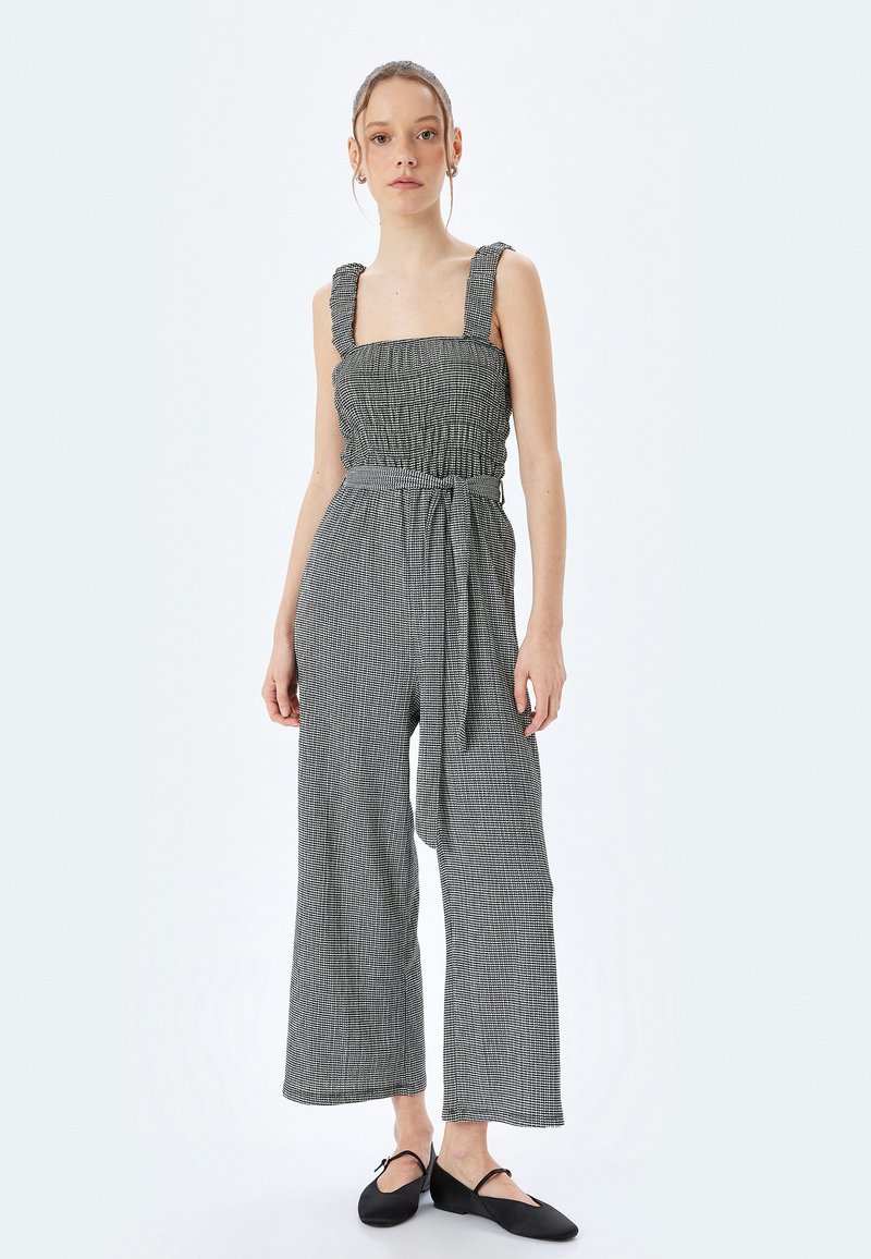 Jumpsuit a righe bianche e nere con gambe larghe, corpetto arricciato e spalline spesse. Include una cintura in tessuto per una vestibilità regolabile.