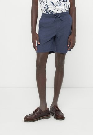 Granatowe spodenki do kolan z elastycznym pasem i sznurkiem, w połączeniu z brązowymi skórzanymi butami żeglarskimi.