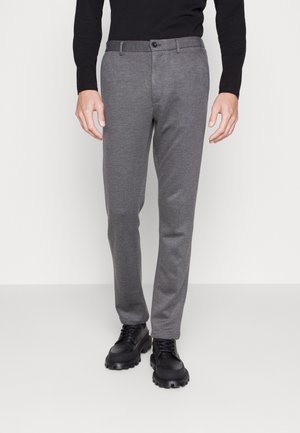 Uomo che indossa pantaloni grigi aderenti, scarpe nere robuste con lacci e una maglia nera a maniche lunghe, in piedi davanti a uno sfondo semplice.