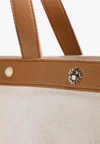 Canvas draagtas met een lichtbruine textuur van leren handvatten, gouden zon-embleem en klinknageldetails. Heeft een lichte, stevige stoffen body.