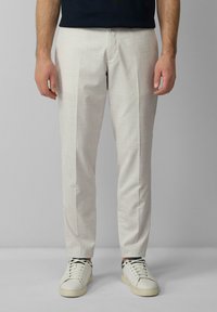 Helle beige, maßgeschneiderte Hose mit strukturiertem Gewebe, die eine schmale Passform und einen gebügelten Frontschnitt aufweist. Kombiniert mit weißen Sneakers und einem dunklen Oberteil.