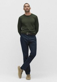 Vihreä neulepusero pyöreällä pääntiellä, tummansiniset housut, letitetty vyö ja vaaleat tennarit. Yksinkertainen muotoilu, sileä tekstuuri ja neutraali väriharmonia.