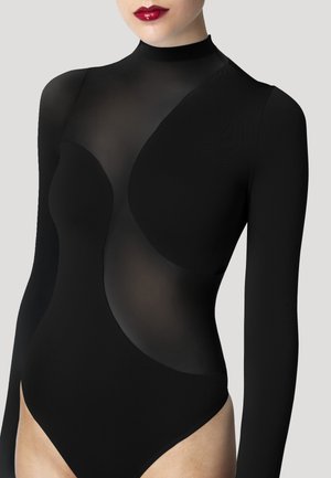 Kvinde iført en sort gennemsigtig heldragt med uigennemsigtige paneler og lange ærmer, læber malet med glinsende rød læbestift, foran en ensartet lys baggrund.