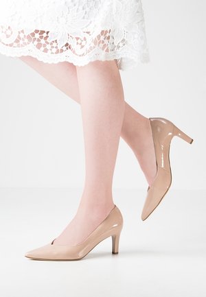 Nude z lakierowanej skóry wysokie obcasy z ostrym noskiem, 5 cm obcasem szpilką i gładkim, błyszczącym wykończeniem, zestawione z białą koronkową sukienką.