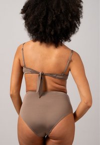 Hochgeschnittener Bikini aus leicht braunem Stoff. Das Oberteil hat dünne, verstellbare Träger und ein Bindeband-Design. Glatte Textur, minimales Zubehör.