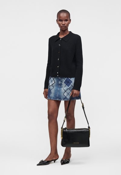Femme portant un cardigan noir boutonné, une jupe en jean bleue à motifs, des talons noirs pointus, et tenant un sac à main en cuir noir.