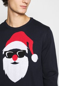 Mörkblå stickad tröja med en julgran-motiv av jultomte med röd hatt, svarta solglasögon och vit skägg. Mjuk textur, rundad halsringning.