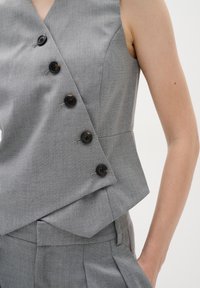 Grau ärmelloser Blazer mit Wickeldesign, ausgestattet mit großen schwarzen Knöpfen, strukturiertem Stoff und tailliertem Schnitt, kombiniert mit passender Hose.
