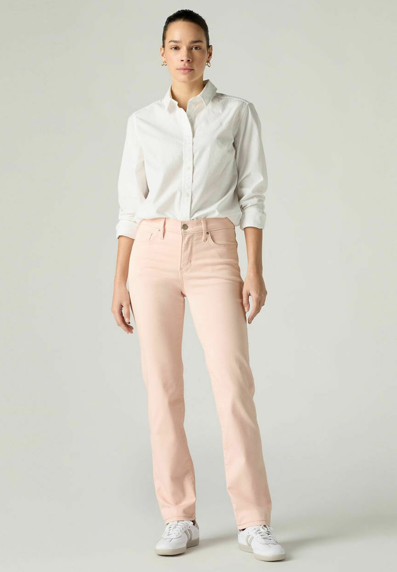 Jean rose clair à coupe slim, associé à une chemise blanche boutonnée. La tenue présente un design simple et est complétée par des baskets blanches.