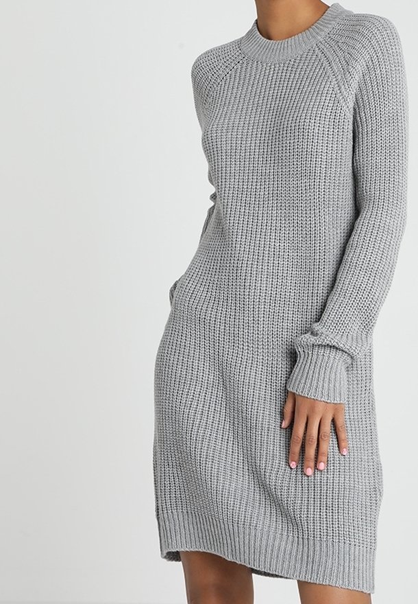 Femme portant une robe pull en maille grise à manches longues, longueur genoux, avec poignets et ourlet côtelés, debout devant un fond uni.