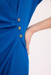 Niebieska sukienka z gładkiego materiału, z wywiniętym detalem w pasie i czterema złotymi guzikami w kształcie róży z boku.