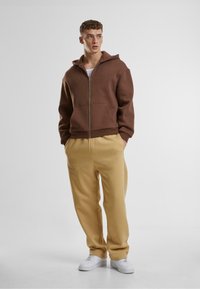 Brauner Kapuzenpulli mit Reißverschluss, kombiniert mit beigen Jogginghosen und weißen Sneakers. Einfaches Design mit einer entspannten Passform und minimalen Akzenten.