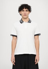 Camisa polo branca com colarinho com padrão de zebra, detalhes pretos nas mangas e "VERSACE JEANS COUTURE" bordado no peito.