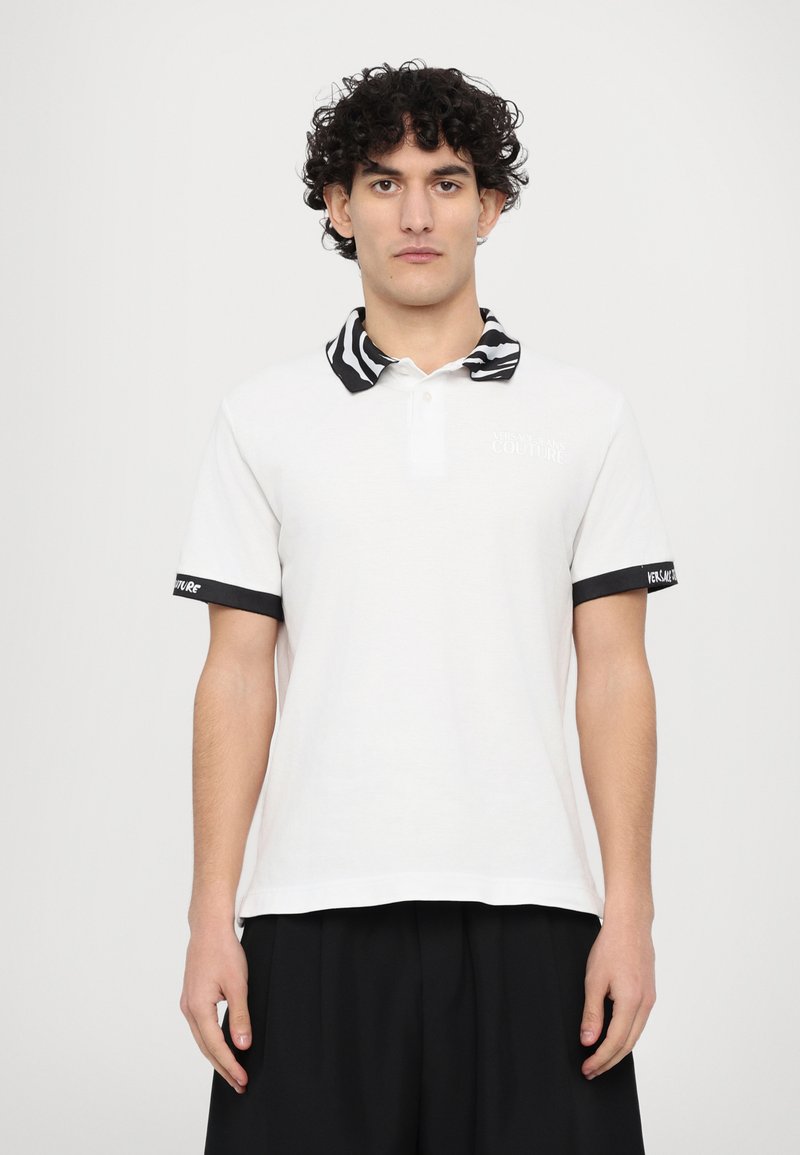 Camisa polo branca com colarinho com padrão de zebra, detalhes pretos nas mangas e "VERSACE JEANS COUTURE" bordado no peito.