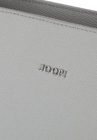 Szary materiał o teksturze z gładkim wykończeniem, z wyraźnie umieszczonym srebrnym tłoczonym logo "JOOP!".