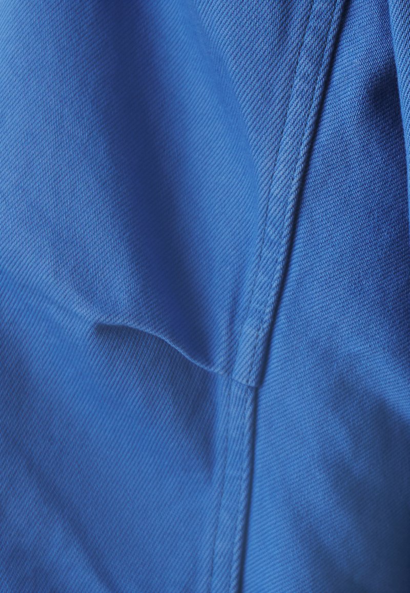 Blauer Stoff mit strukturierter Oberfläche, der glatte und gerippte Muster aufweist sowie sichtbare Nähte. Nahaufnahme hebt die Materialdetails hervor.