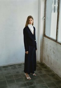 Námornícky kardigán so zlatým gombíkom, nosený cez dlhú námornícku sukňu. Biela šatka pridáva kontrast. Model stojí v minimalistickom, dobre osvetlenom priestore.