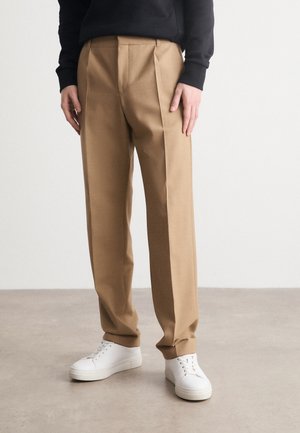 Tan pleated trousers made of woven fabric, featuring a straight leg design. Paired with white sneakers on a neutral surface.