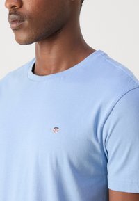Light blue cotton t-shirt featuring a round neck, short sleeves, and a small embroidered logo with a shield design on the chest.