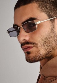 Ochelari de soare dreptunghiulari cu lentile întunecate, rame din metal argintiu și accente cu logo. Temple texturate și design elegant.