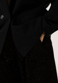 Blazer noir avec des revers crantés et deux boutons, associé à un pantalon sombre et texturé arborant un motif de points dorés dispersés.