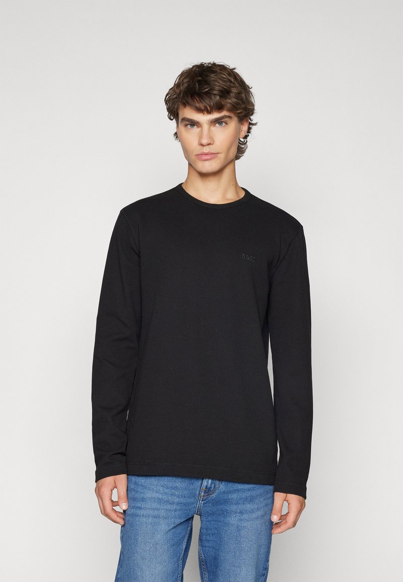 Zwarte longsleeve T-shirt met een ronde hals, gemaakt van ribbelstof. Heeft subtiele branding op de borst, gecombineerd met blauwe jeans.