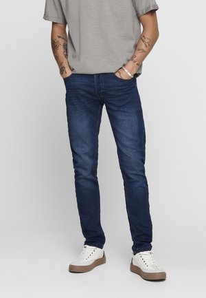 Slim-fit dark denim jeans with slight fading, featuring five pockets and a zip fly. Worn with a gray t-shirt and white sneakers.