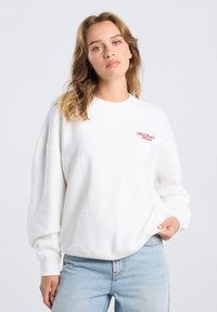 Sudadera blanca de manga larga, con puños acanalados, cuello redondo y texto bordado en rojo que dice "New York City". Textura de tejido suave.