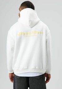 Sweat-shirt à capuche blanc avec texte brodé jaune "About: Vibes" et "Crafted Rebellion" au dos ; doté d'une texture douce et d'un ourlet côtelé.