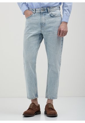 Osoba ubrana w jasnoniebieskie wyprane dżinsy, brązowe sznurowane buty i niebieską koszulę w paski, stojąca z jedną ręką w kieszeni.