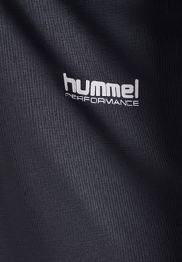 Zbliżenie na czarną teksturowaną tkaninę z białym logo "hummel PERFORMANCE" wydrukowanym na powierzchni.
