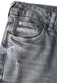 Gros plan sur une poche de jean en denim bleu clair avec des rivets en métal et une petite déchirure usée en dessous de la poche.