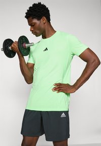 Groene sport T-shirt met zwart logo, gedragen met donkere shorts. Persoon houdt een dumbbell vast, wat een gespierd uiterlijk en een gefocuste houding laat zien.