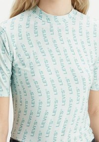 Světlé modré tričko s krátkým rukávem s vzorem "LEVI'S" v různých orientacích. Hladká textura a jednoduchý design s kulatým výstřihem.