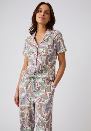 Femme portant une chemise courte à boutons et un pantalon imprimé de feuilles tropicales vertes, roses et violettes, posant avec une main dans la poche.