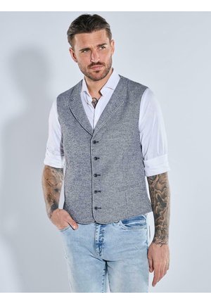 Mann mit Tätowierungen, der eine graue, geknöpfte Weste über einem weißen, hochgekrempelten Hemd und hellblauen Jeans trägt, steht vor einem neutralen Hintergrund.