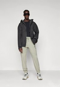 Vannavstøtende svart jakke med justerbar hette, brukt over en mørk topp og lysebeige avsmalnende bukser, komplettert med store sneakers.