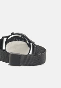 Černé hodinky se stahovacím nerezovým páskem. Mají kulatý ciferník a průhledné sklo na zadní straně. Minimalistický design s bezpečným uzavíráním.