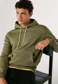 Olijfgroene hoodie met een voorzak, witte koorden en geribde manchetten. Draag ik over een gele shirt, gecombineerd met zwarte broek.