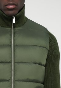 Roheline puffer vest kõrge kraega ja hõbedase tõmblukuga. Tekstuuriga kudumihari varrukatel. Sileda, polsterdatud lõikega elemendid loovad struktureeritud välimuse.