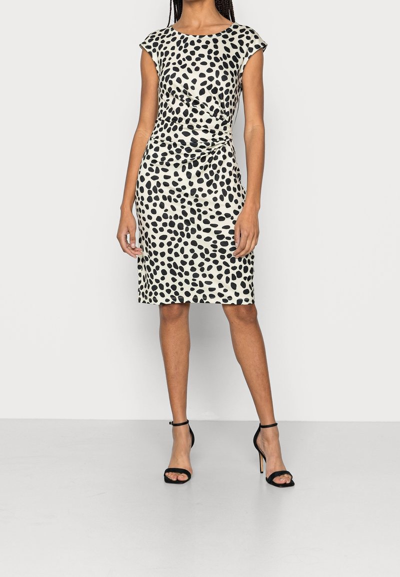 Knieum lange Kleid in Creme mit schwarzem abstraktem Muster, kurzen Ärmeln und einer figurbetonten Silhouette mit einem seitlichen Twist-Detail. Schwarze Sandalen mit Absatz.