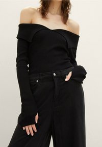 Femme portant un haut noir côtelé à manches longues et épaules dénudées, ainsi qu'un pantalon noir taille haute, une main dans la poche, sur un fond uni.