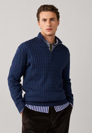 Homme portant un pull à fermeture éclair bleu torsadé sur une chemise à rayures avec un pantalon marron, debout devant un fond uni clair.