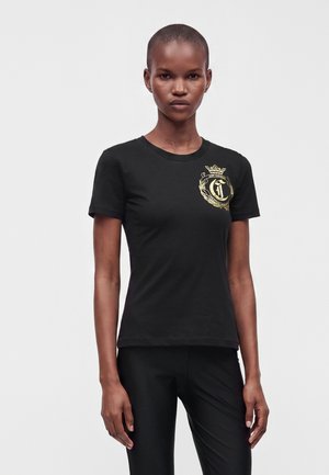 Modelo delgado con cabello corto que lleva una camiseta negra con un emblema de un escudo dorado en el pecho izquierdo y pantalones negros, de pie sobre un fondo blanco.