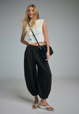 Jeune femme aux longs cheveux blonds portant un top court blanc sans manches, un pantalon noir ample, des sandales compensées blanches et un sac noir porté en bandoulière.